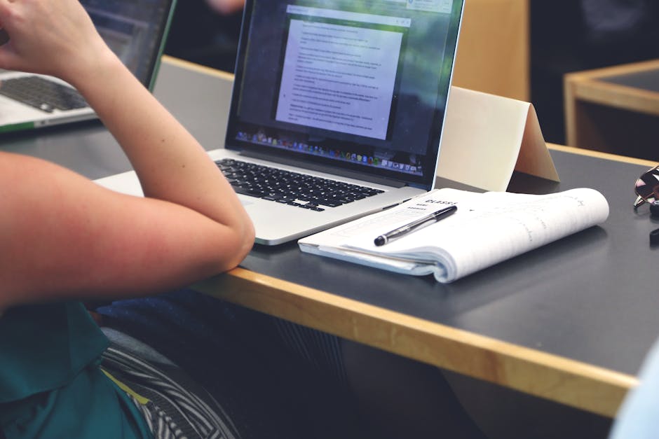 A student in a classroom setting using a laptop and taking notes in a notebook
